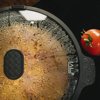 Glasdeckel der Schmorpfanne Almar mit Dampfauslass und Kondenswasser über einer brutzelnden Pfanne, daneben eine frische Tomate auf dunkler Arbeitsplatte.