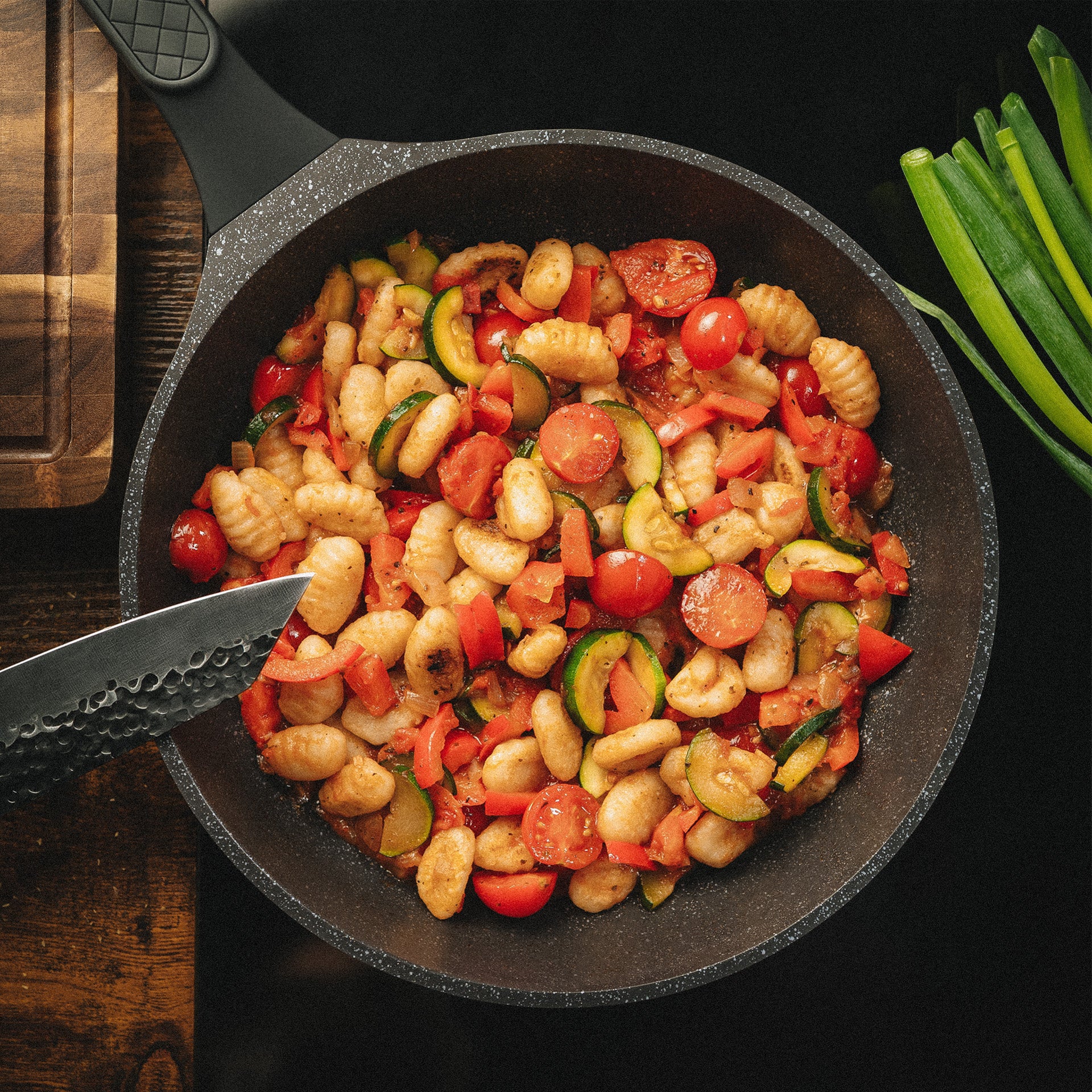 Große Bratpfanne Vanessa mit fertigem Gnocchi-Gericht auf einem schwarzen Kochfeld neben frischen Kräutern.