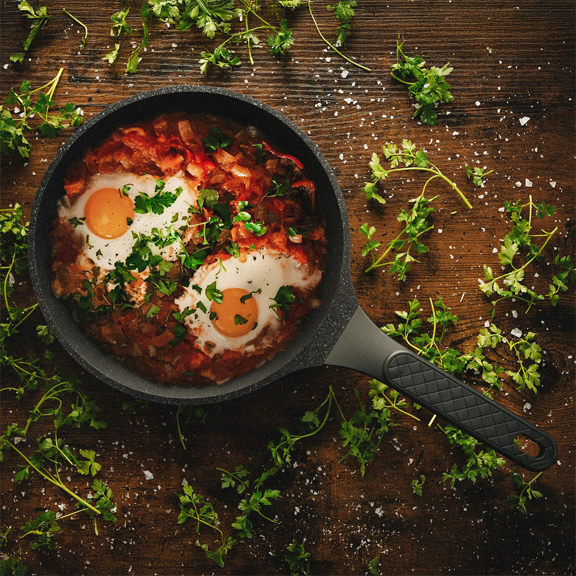 Shakshuka mit pochierten Eiern und frischer Petersilie in einer schwarzen Gussaluminium-Pfanne auf Holzhintergrund.