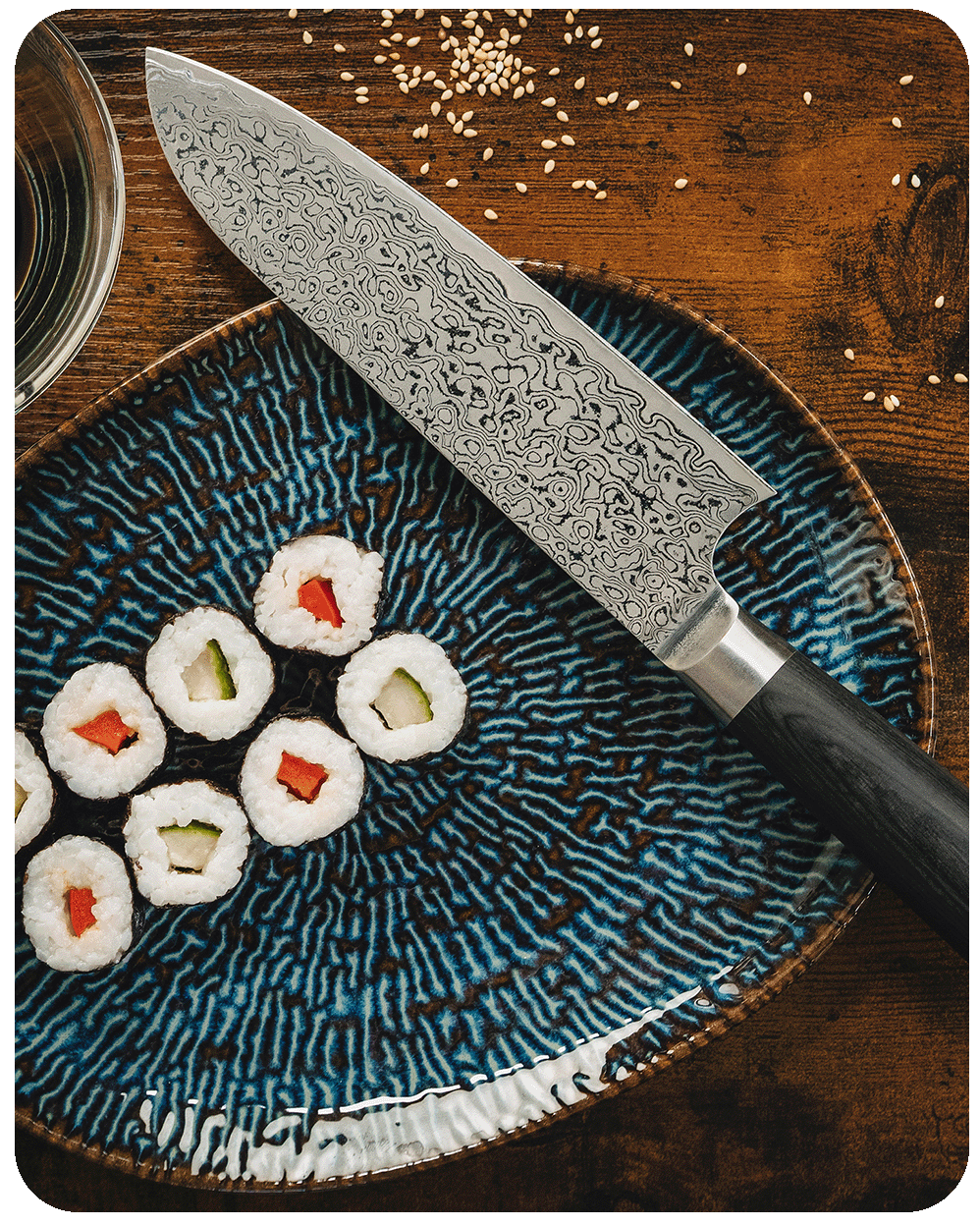 Japanisches Damastmesser neben Sushi-Rollen auf einem blauen Keramikteller.