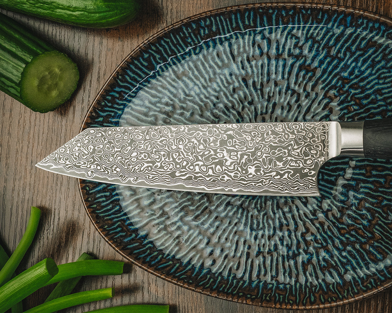 Kiritsuke-Damastmesser flach liegend auf einem gemusterten Teller mit Gemüse-Garnitur.