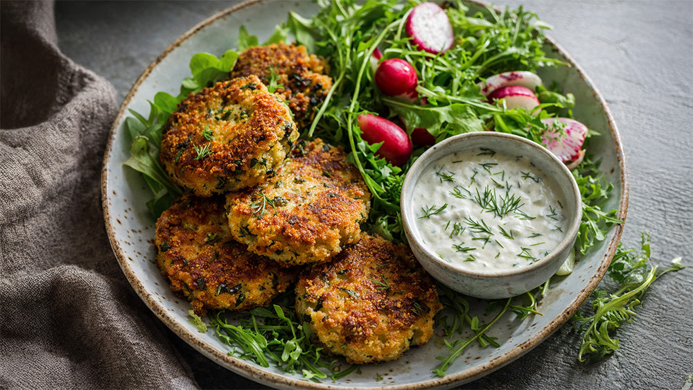 Nahaufnahme von knusprigen Gemüsefrikadellen mit Kräuter-Dip und knackigem Rucola-Salat, serviert auf einem hellen Keramikteller.