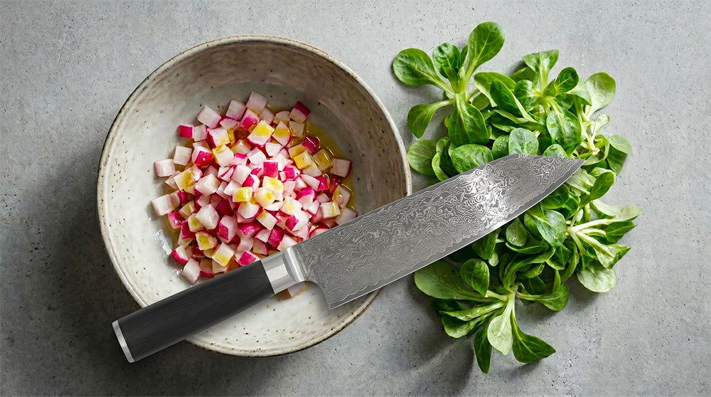 Draufsicht auf eine rustikale Schale mit fein gewürfelten Radieschen in Vinaigrette. Daneben liegt ein Damast-Kochmesser mit dunklem Griff und ein Büschel Feldsalat auf grauem Beton.