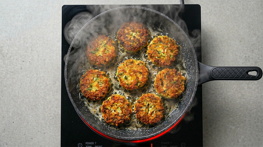 Draufsicht auf eine FIVI Bratpfanne auf einem schwarzen Induktionskochfeld. In der Bratpfanne liegen golden-braun gebratene Gemüsebratlinge und brutzeln vor sich hin.