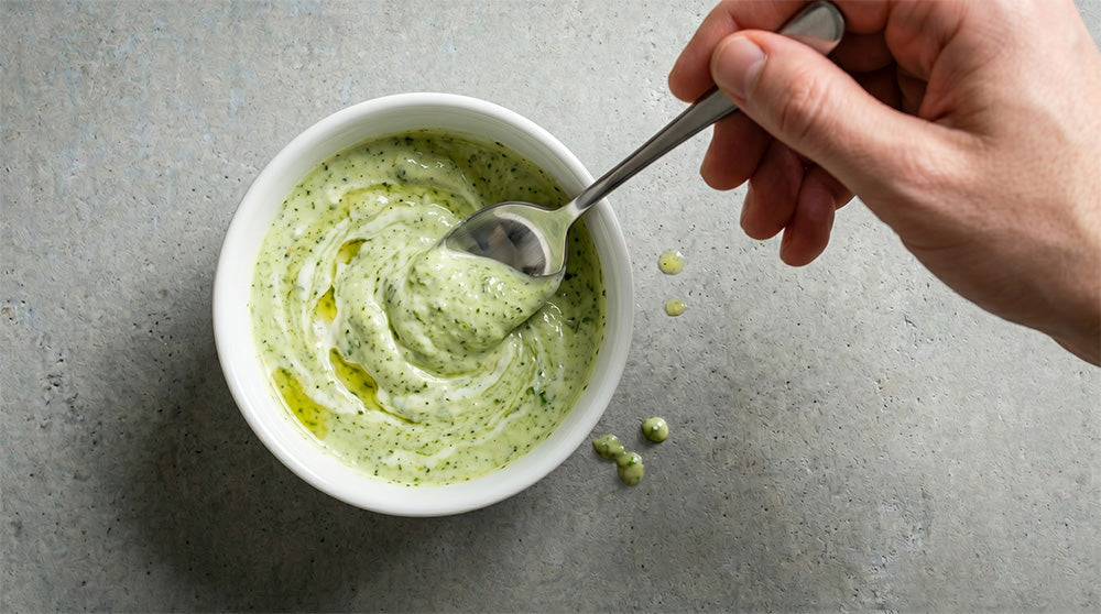 Eine Hand rührt mit einem Löffel in einer weißen Schale mit cremiger Joghurt-Pesto-Sauce, die Olivenöltropfen zeigt. Auf einer grauen Arbeitsfläche.