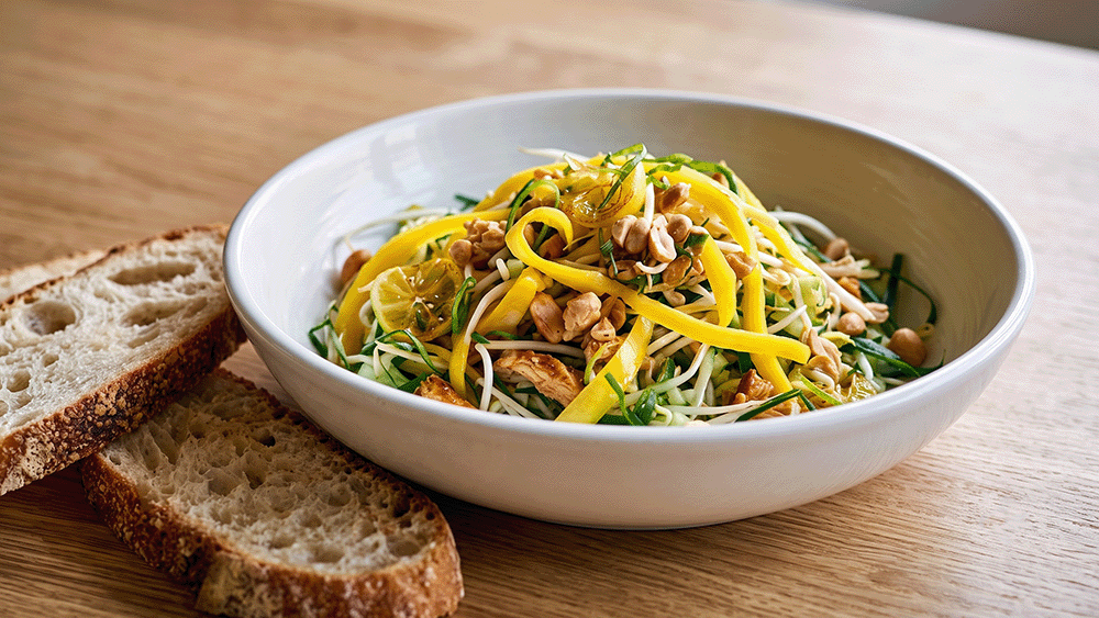 Schräge Nahaufnahme eines bunten Hähnchensalats mit Erdnüssen und Mangostreifen, serviert mit Krustenbrot auf einer hellen Holzoberfläche.