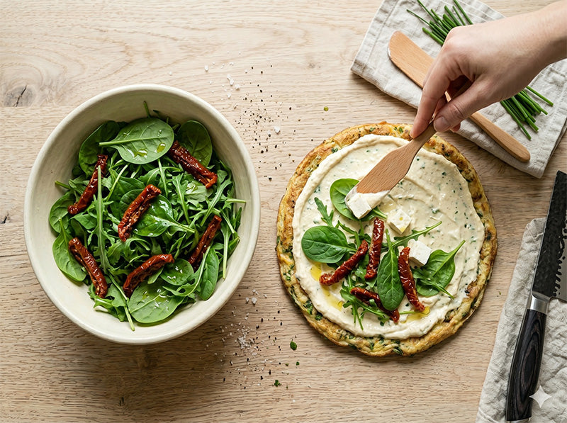 Bestreichen eines Kräuterpfannkuchens mit Feta-Creme, daneben eine Schüssel mit Spinat und getrockneten Tomaten.