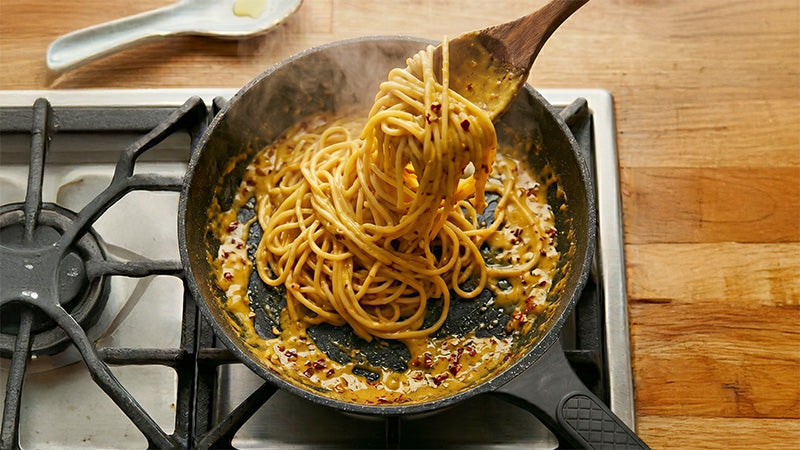 Spaghetti werden in der Pfanne mit glänzender, goldener Maissauce und Chiliflocken vermengt.