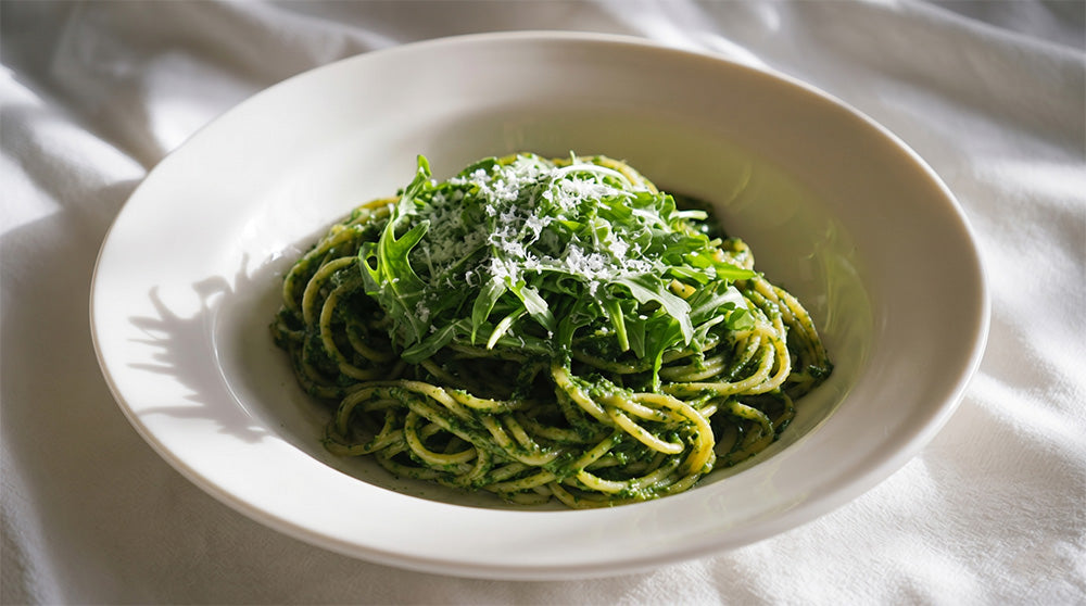 Schräge Nahaufnahme von Spaghetti in grüner Kräutersauce, getoppt mit Rucola und Parmesan in einem hellen Teller bei natürlichem Lichteinfall.