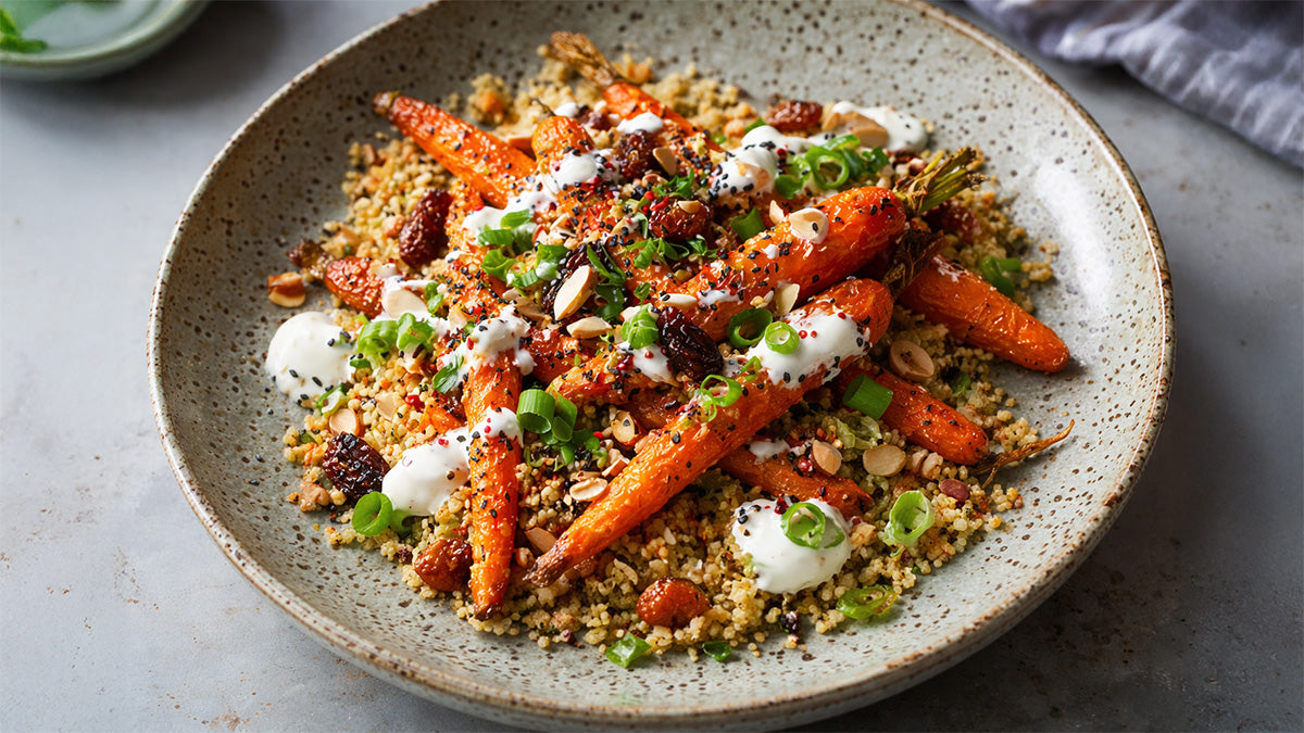 Nahaufnahme von gerösteten Möhren mit Raz-el-Hanout auf Couscous. Die glänzenden, gewürzten Karotten sind mit weißem Joghurt-Dressing, Sesam und Kräutern getoppt. Ein stimmungsvolles Food-Foto in warmem Licht.