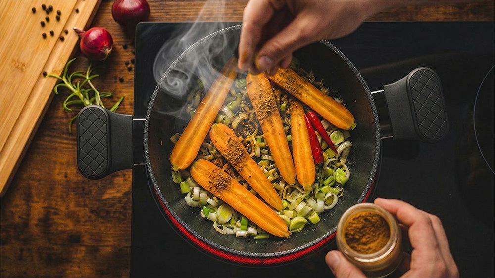 Halbiert Möhren und Frühlingszwiebeln werden in einem dunklen Topf auf einem Induktionsherd angebraten. Eine Hand bestreut das Gemüse mit Raz el Hanout Gewürz, während leichter Dampf aufsteigt.