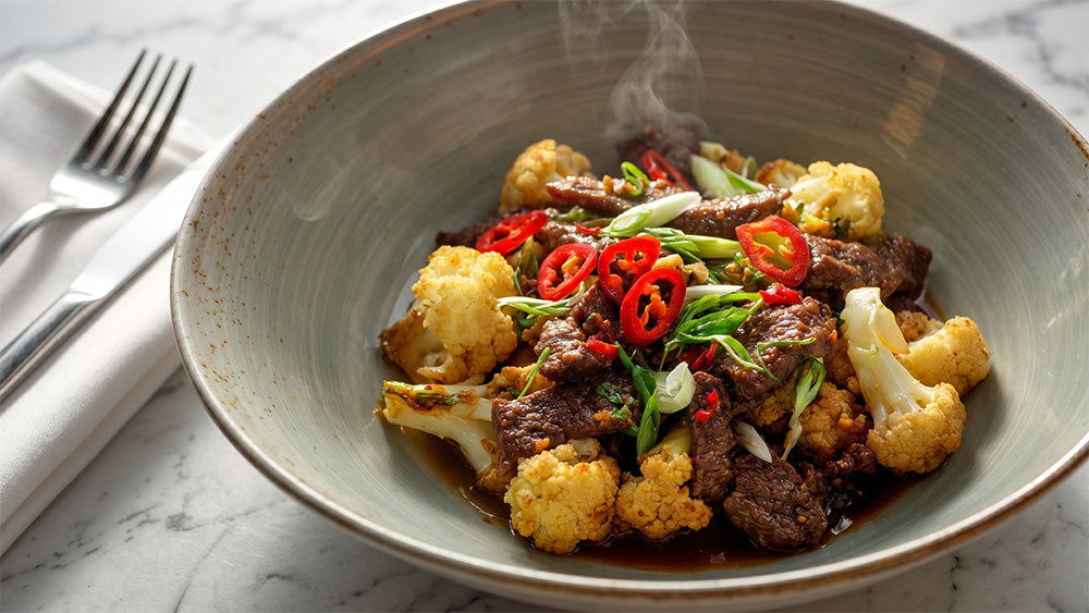 Nahaufnahme aus schräger Perspektive von dampfendem Rindfleisch-Blumenkohl-Stir-Fry mit Chili in einer rustikalen Keramikschüssel.