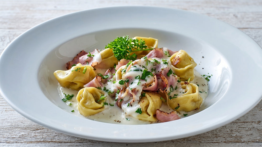 Nahaufnahme von Tortellini in Sahnesauce mit krossen Schinkenstreifen und frischer Petersilie auf hellem Holzgrund.